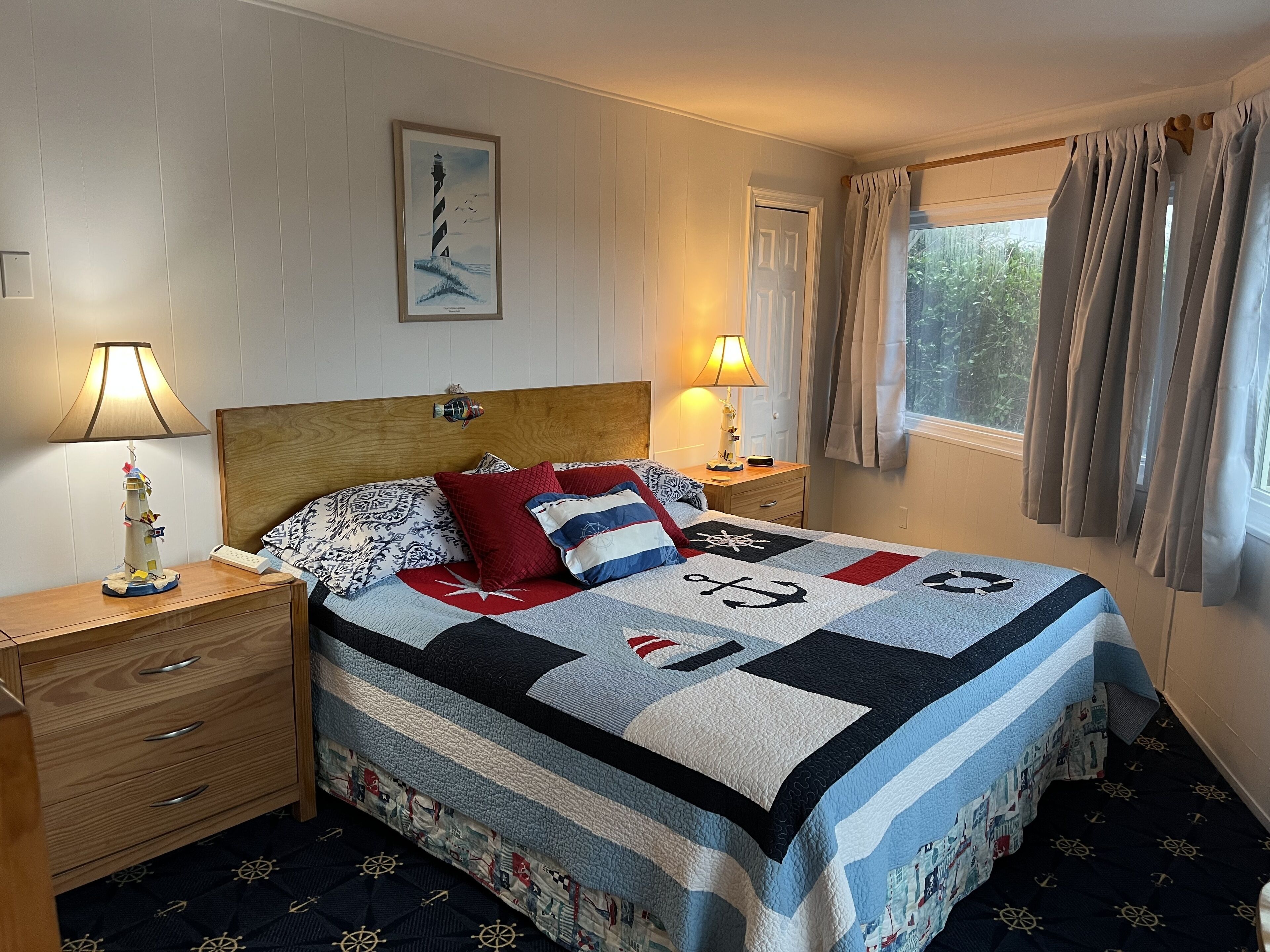 A neatly made bed with a quilt featuring nautical designs, two bedside lamps, a wooden nightstand with drawers, and a window with curtains.