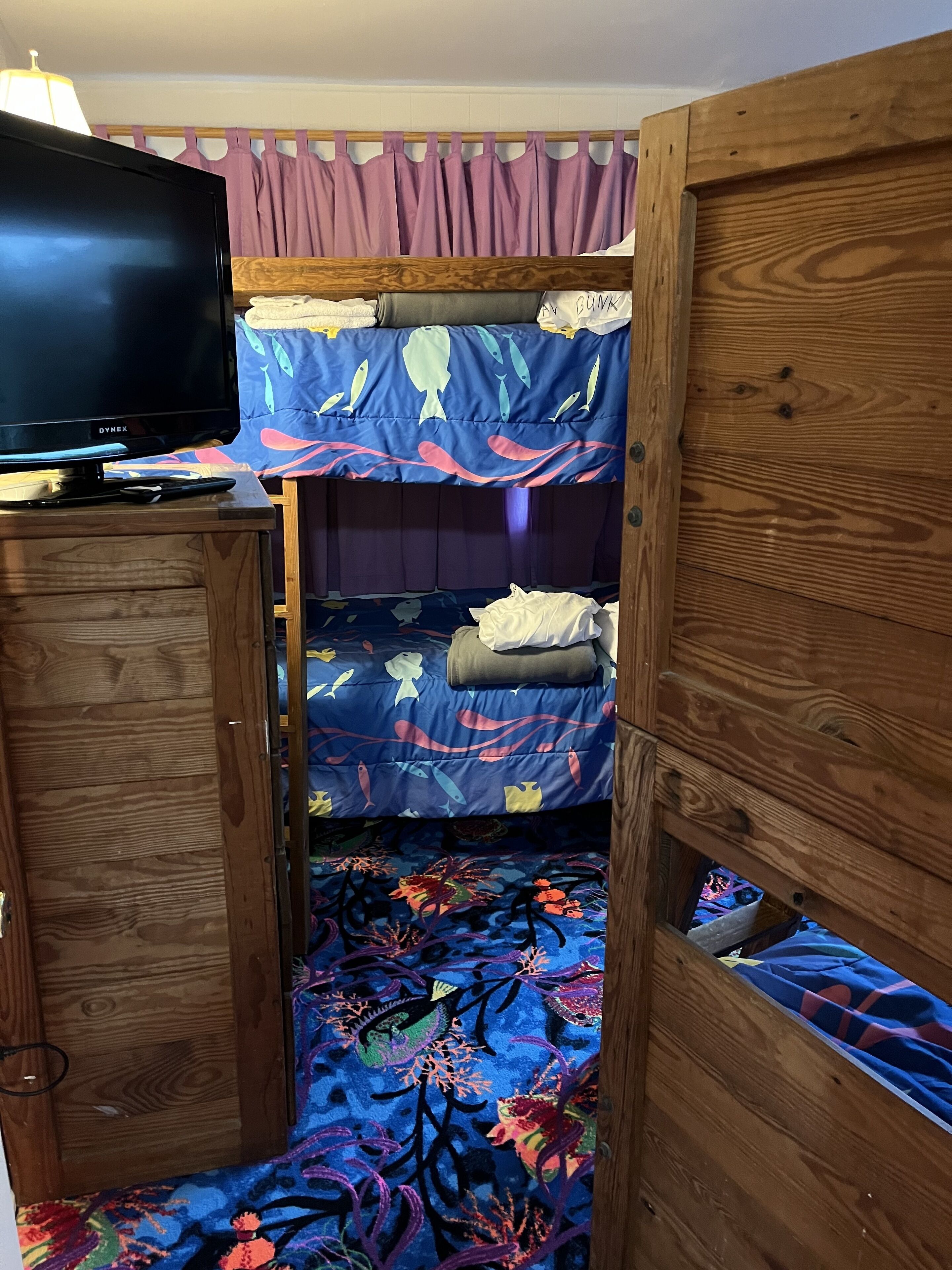 A bunk bed room with a television on a wooden cabinet, a purple curtain, and a bed with a blue and red fish pattern.