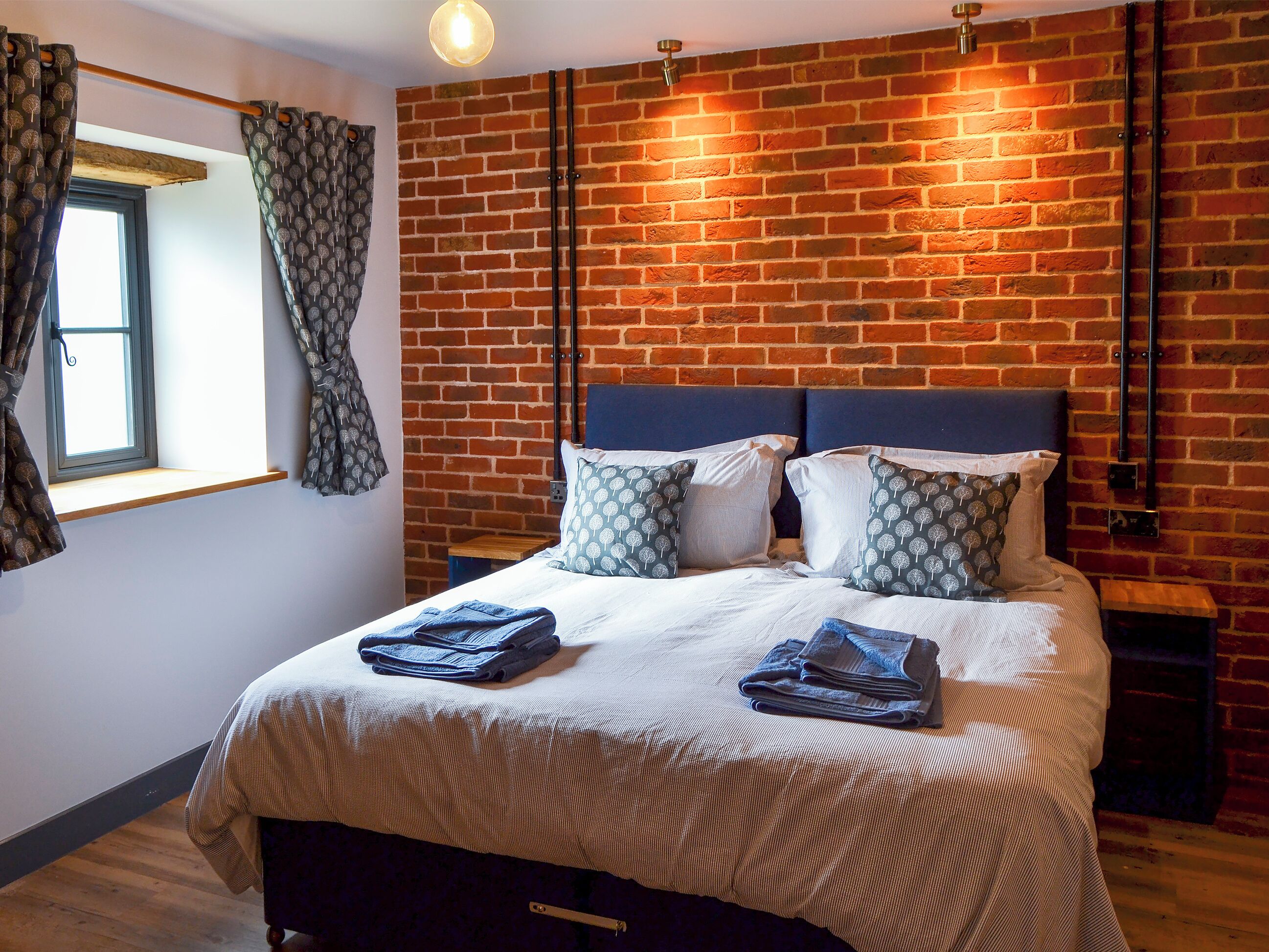 Une chambre avec un mur en briques, un lit avec une tête de lit, une fenêtre avec des rideaux et une table de chevet.