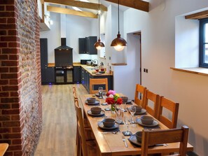 A dining area with a wooden table set for six, flanked by brick walls and wooden beams. A kitchen with dark cabinetry and a chimney is visible in the background.