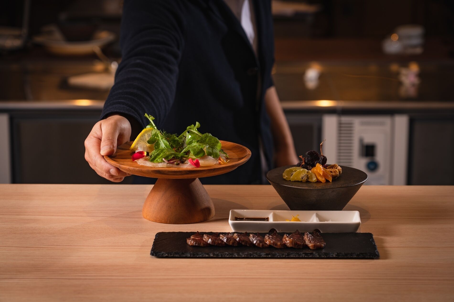 A person serving a dish with greens and a bowl of dried fruits on a wooden table.