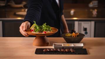 A person serving a dish with greens and a bowl of dried fruits on a wooden table.