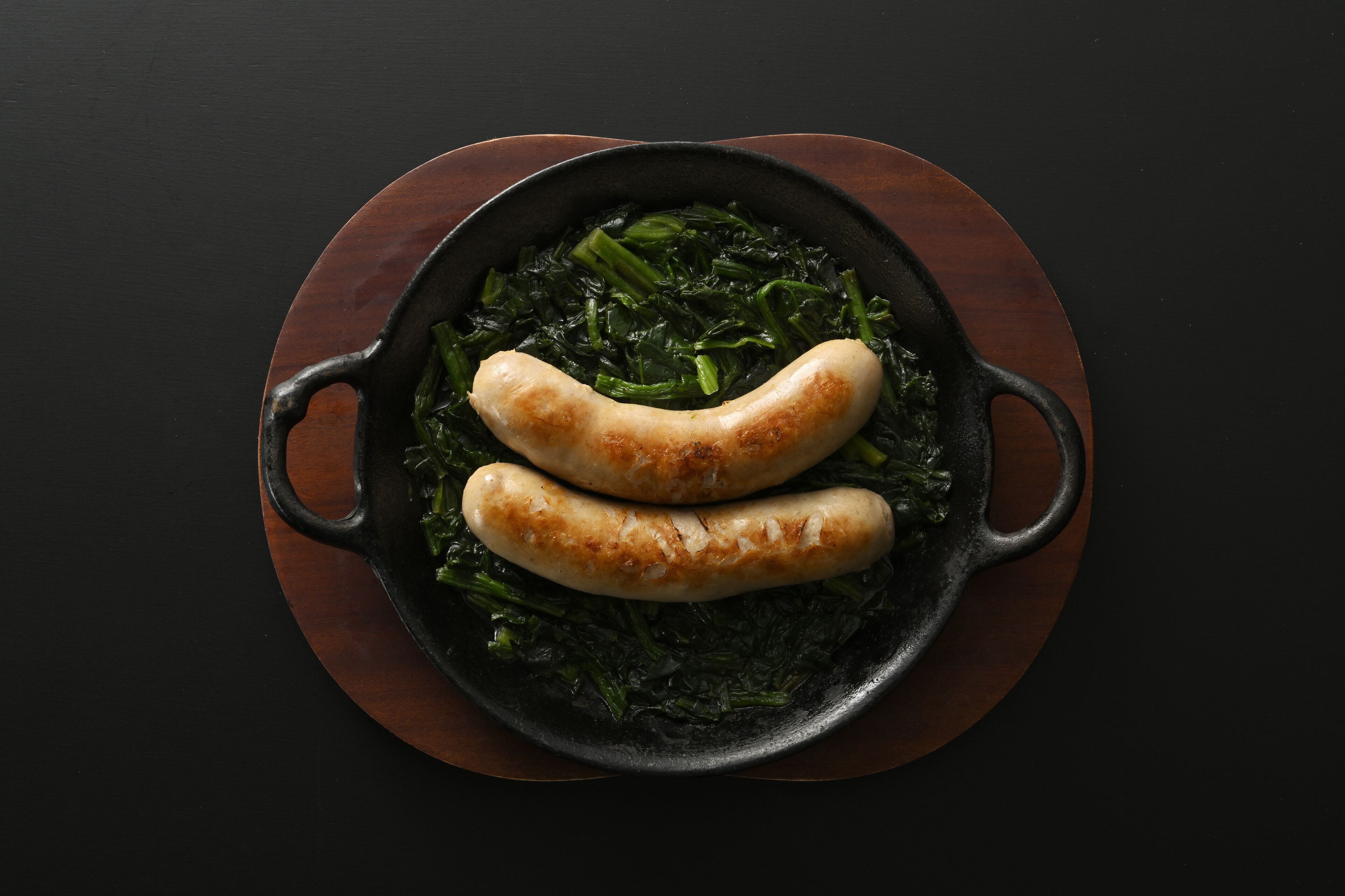 Una sartén con dos salchichas y vegetales sobre una tabla de madera.