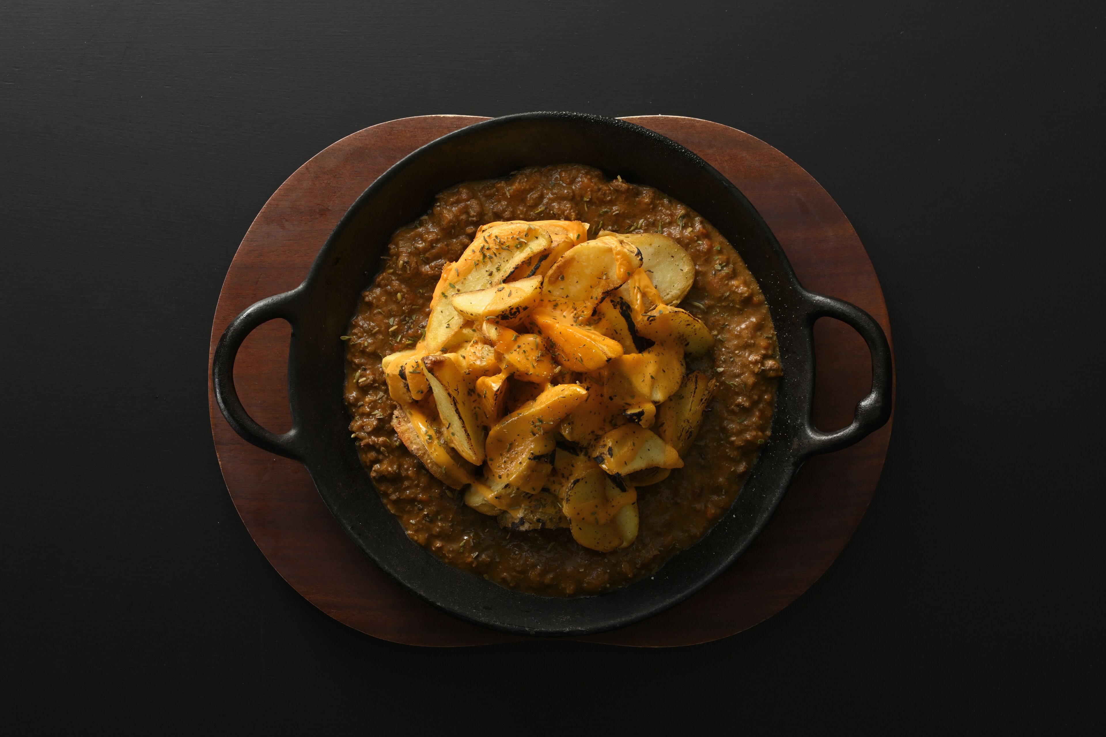 A skillet with a stew-like dish and sliced potatoes on a wooden board.