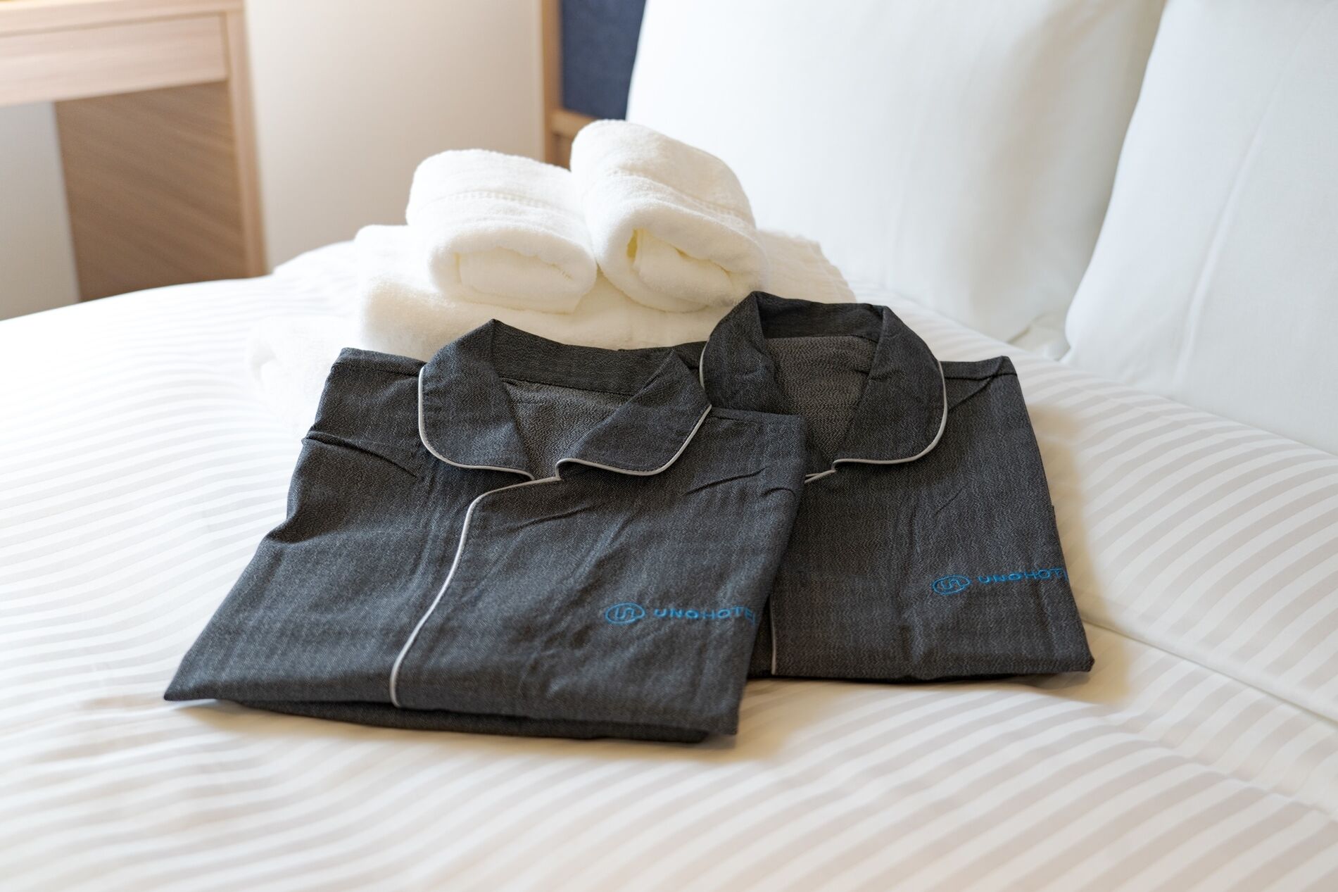 A neatly made bed with a white pillow and two folded dark grey shirts with a collar and button placket, and a white towel.