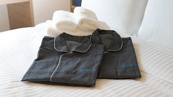 A neatly made bed with a white pillow and two folded dark grey shirts with a collar and button placket, and a white towel.