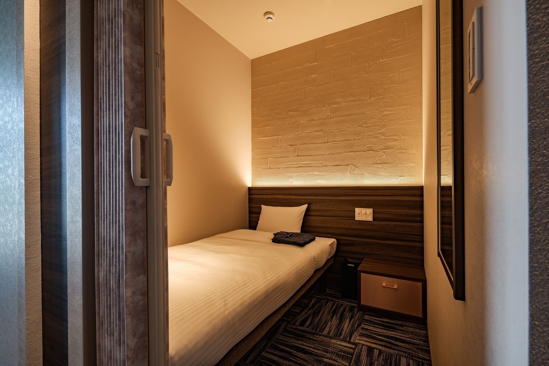 A compact hotel room with a single bed, a small bedside table, a nightstand, a wall-mounted TV, and a window with blinds.
