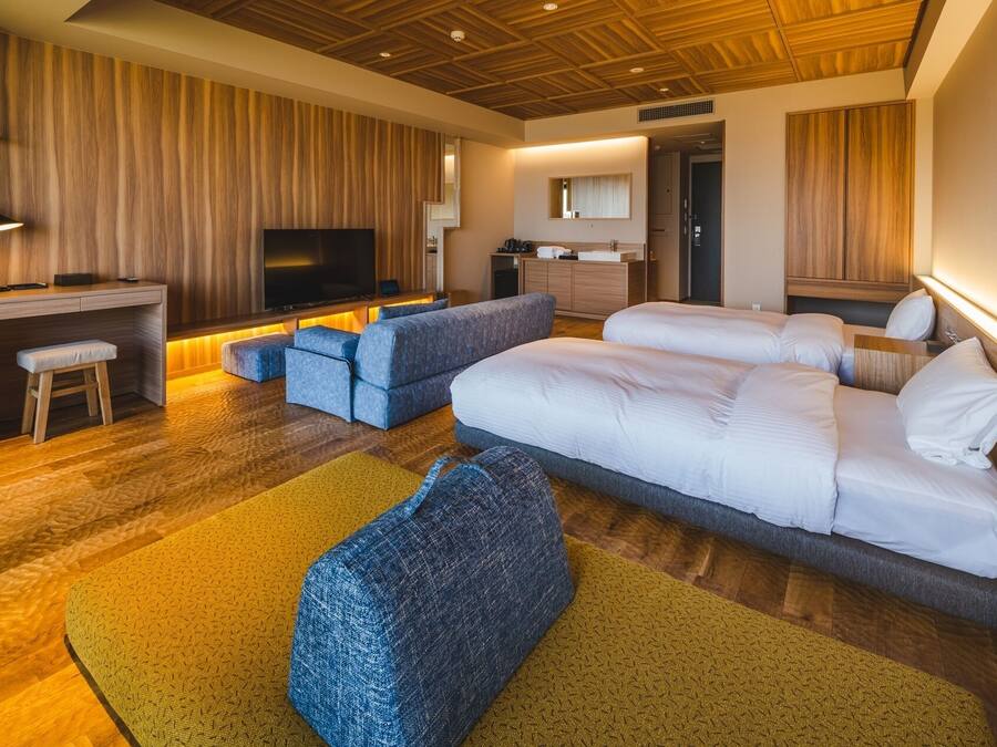 Een moderne hotelkamer met een houten plafond, een flatscreen televisie, een bureau met stoel, twee bedden en een zithoek met poefjes.