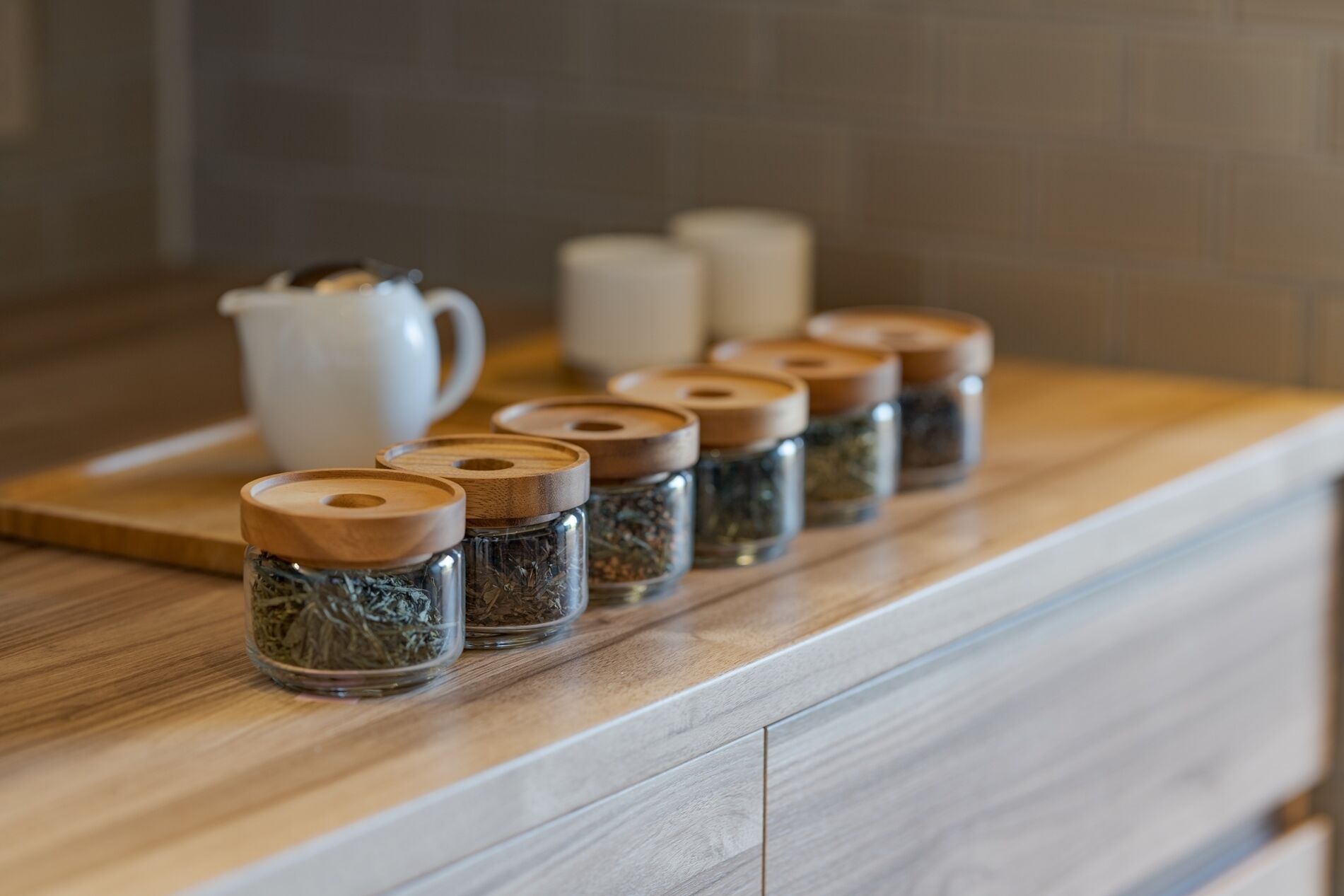 A kitchen counter with several glass containers with wooden lids, a white pitcher, and a white container in the background.
