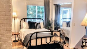 Ein Schlafzimmer mit einem Bett, einem Deckenventilator, einem Fenster mit Vorhängen, einem Nachttisch und einem Beistelltisch.