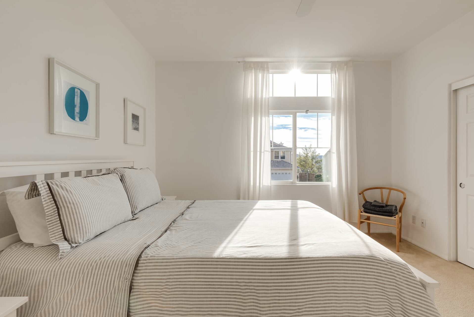 A bedroom with a large bed, a window with curtains, a chair, and two framed pictures on the wall.