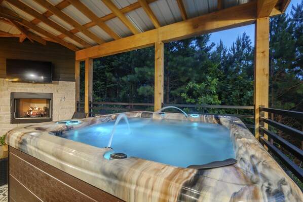 A hot tub with a stone surround and a fireplace, set in an outdoor wooden deck with a view of trees.