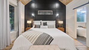 Un dormitorio con una cama grande, dos mesitas de noche con lámparas, un ventilador de techo y vistas al baño.