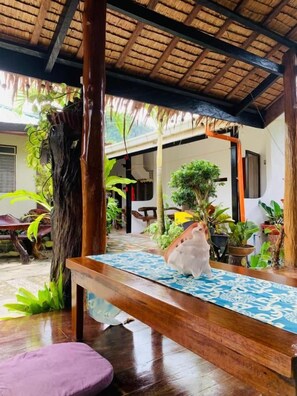 A wooden bench with a patterned cushion, a tree trunk, and potted plants in a covered outdoor area. - Pisces Garden Tourist Inn (San Vicente)