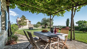 Une terrasse en pierre avec une table et des chaises sous une pergola, entourée de verdure et avec un bâtiment en pierre en arrière-plan.