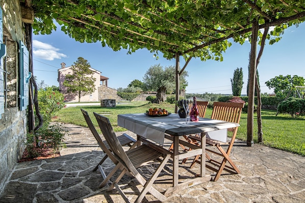 Eine Steinterrasse mit Tisch und Stühlen unter einer Pergola, umgeben von Grünpflanzen und einem Steinhaus im Hintergrund.