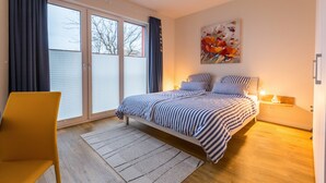 A bedroom with a bed, a chair, a nightstand, a lamp, a painting, and a sliding glass door. - Modern Vacation Apartment "Büsumperle Rot" in Central Location with Wi-Fi (Büsum)