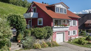 Une maison rouge avec un balcon blanc, un garage et un jardin où poussent différentes sortes de plantes.