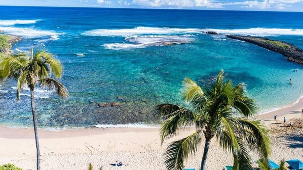 Une plage à l’eau turquoise et limpide, bordée de palmiers et d’une côte rocheuse.