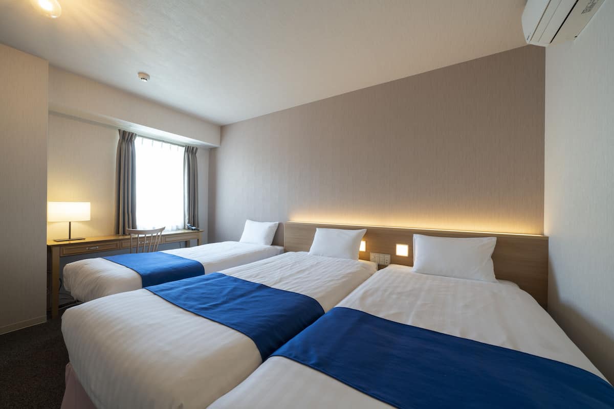 A hotel room with three beds, a desk, a chair, and a window with curtains.