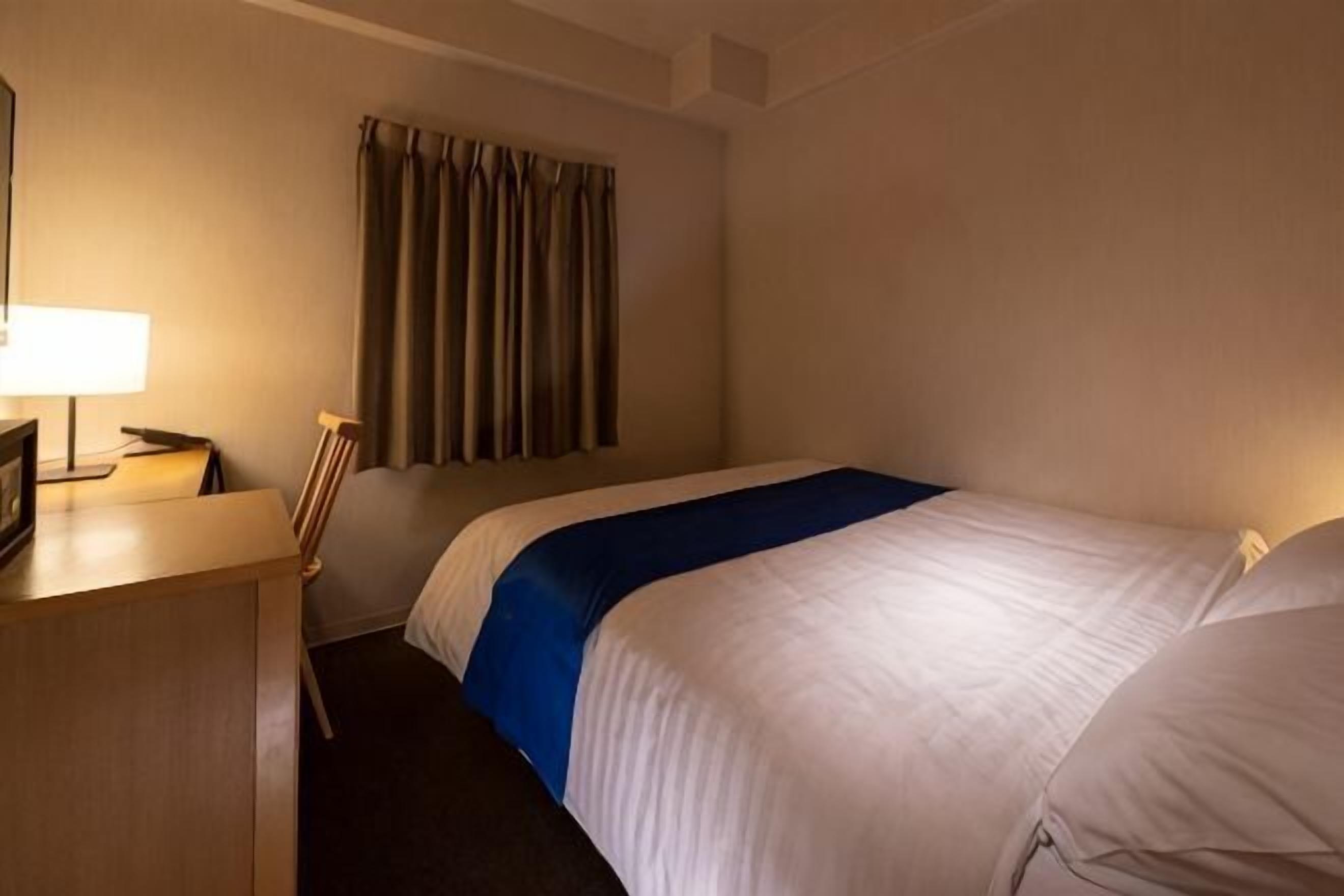 Une chambre d’hôtel avec un lit, un bureau avec une lampe, une chaise et une fenêtre avec des rideaux.