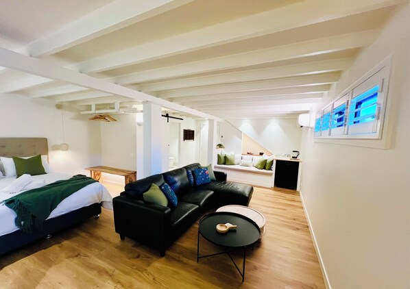 Una habitación moderna y bien iluminada con una cama, un sofá de cuero negro, una pequeña mesa redonda y una zona de cocina al fondo.