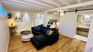 Ein modernes Wohnzimmer mit einer schwarzen Ledersofa, einem runden Couchtisch und einem kleinen Hocker. Der Boden ist aus Holz, die Decke weiß mit sichtbaren Balken, und an der Wand hängt ein Gemälde.