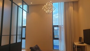 A modern living room with a large window, a chandelier, a desk with a computer, and a sofa. - Hotel Libre (Siheung)