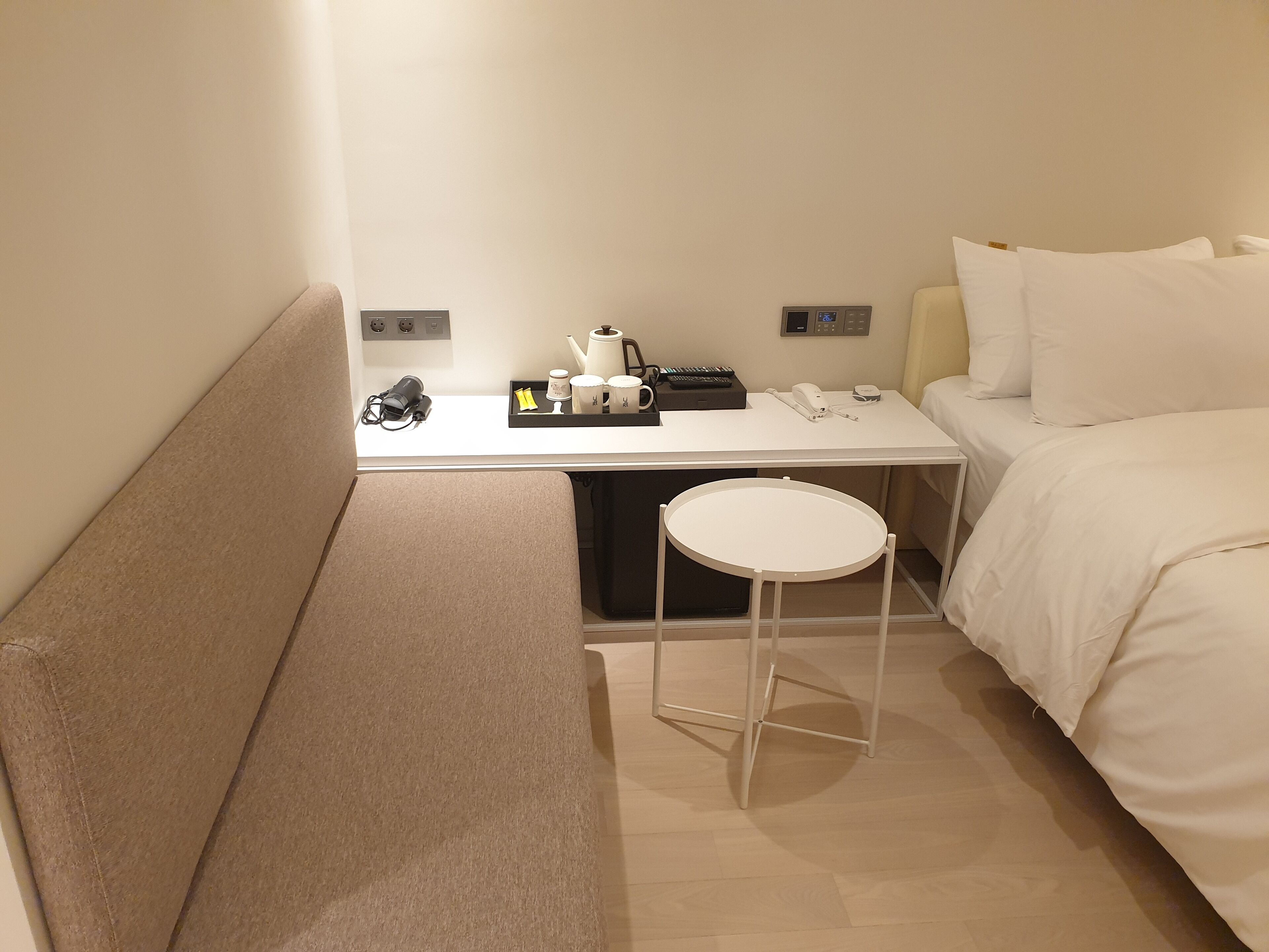 A hotel room with a bed, a small table with a kettle, cups, and a phone. There is a bench and a stool.