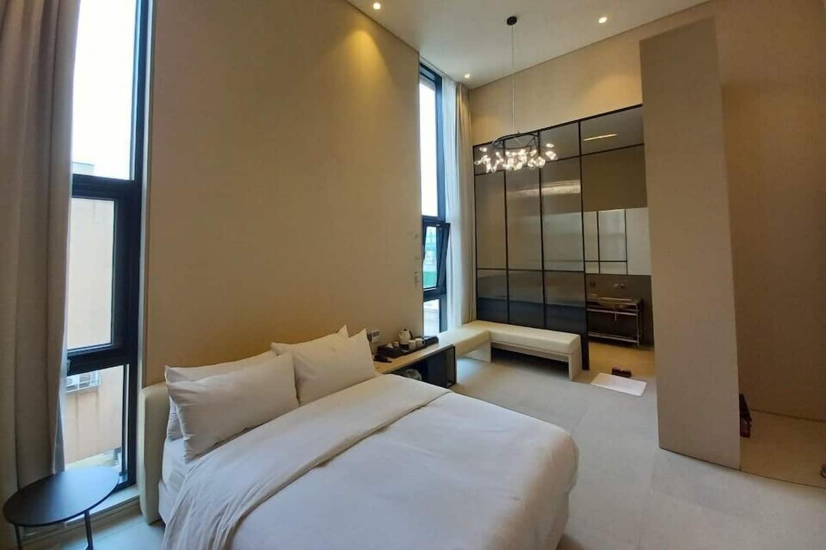 A modern hotel room with a large bed, a small table, a seating area, a large mirror, and a view of the city.