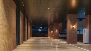 A covered parking area with brick walls and a concrete floor, featuring yellow parking lines and a white utility box. - Hotel Libre (Siheung)