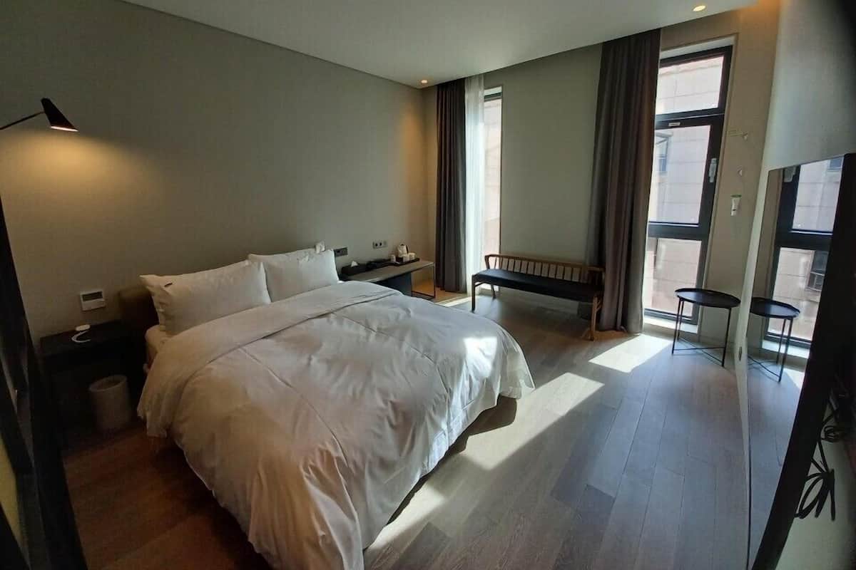 A modern hotel room with a large bed, a bedside table, a bench, and a view of the city through large windows.
