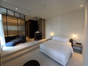 A modern hotel room with a large bed, a seating area with a sofa and a small table, a wardrobe, and a built-in TV. - Hotel Libre (Siheung)