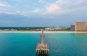 A pier extending into turquoise waters with a beach and buildings in the background.