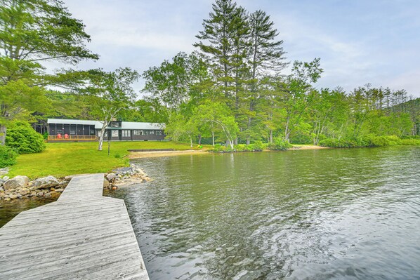 A wooden dock leading to a house with a green roof, surrounded by lush greenery and a calm lake. - Lakefront Escape w/ Deck & Kayaks in Lovell! (Lovell)