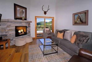 A living room with a stone fireplace, a couch with patterned cushions, a glass coffee table, and a mounted deer head. - Sagebrush, Ski in/out, hot tub, dog friendly (Gunnison)