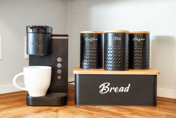 A coffee maker, three containers labeled Coffee, Tea, and Sugar, and a white mug on a wooden table.