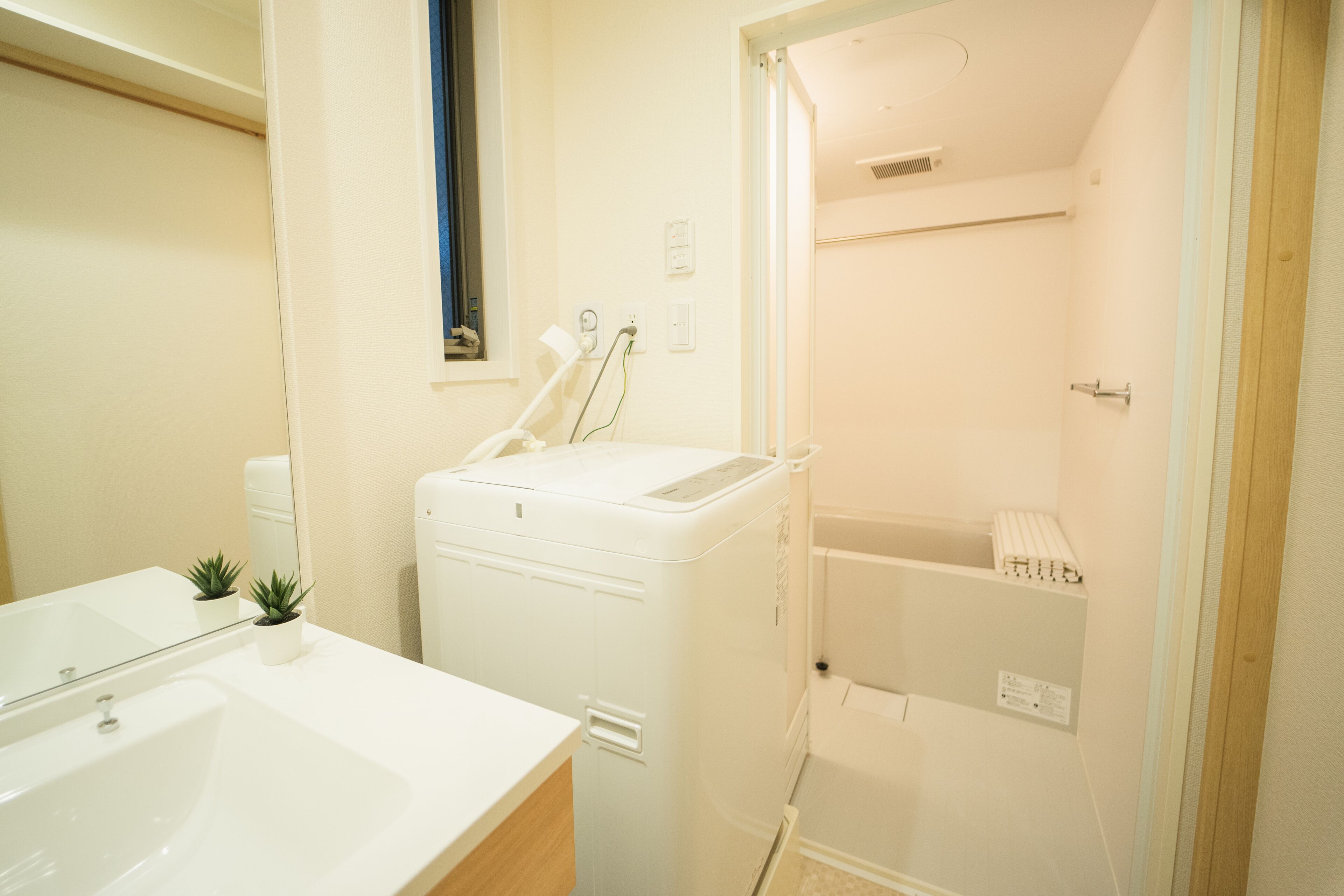 Ein modernes Badezimmer mit einem weißen Waschbecken, einer kompakten Waschmaschine und einem Trockner, einem kleinen Topf mit Pflanze und einer Duschkabine.