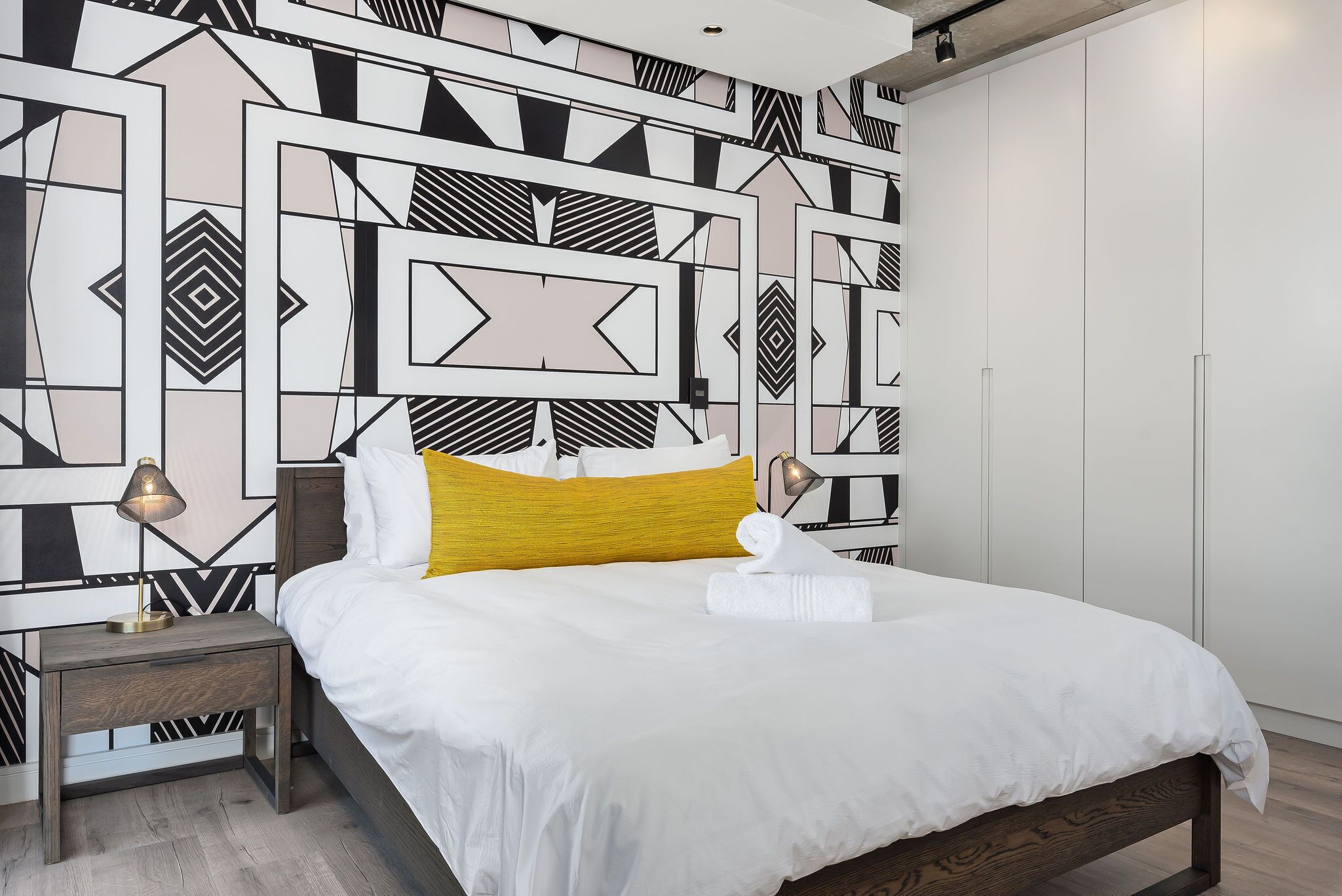 A neatly made bed with white and yellow bedding, a bedside lamp, and a geometric-patterned wall.