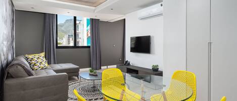 A modern living room with a grey sofa, yellow chairs, a glass coffee table, and a TV on the wall.