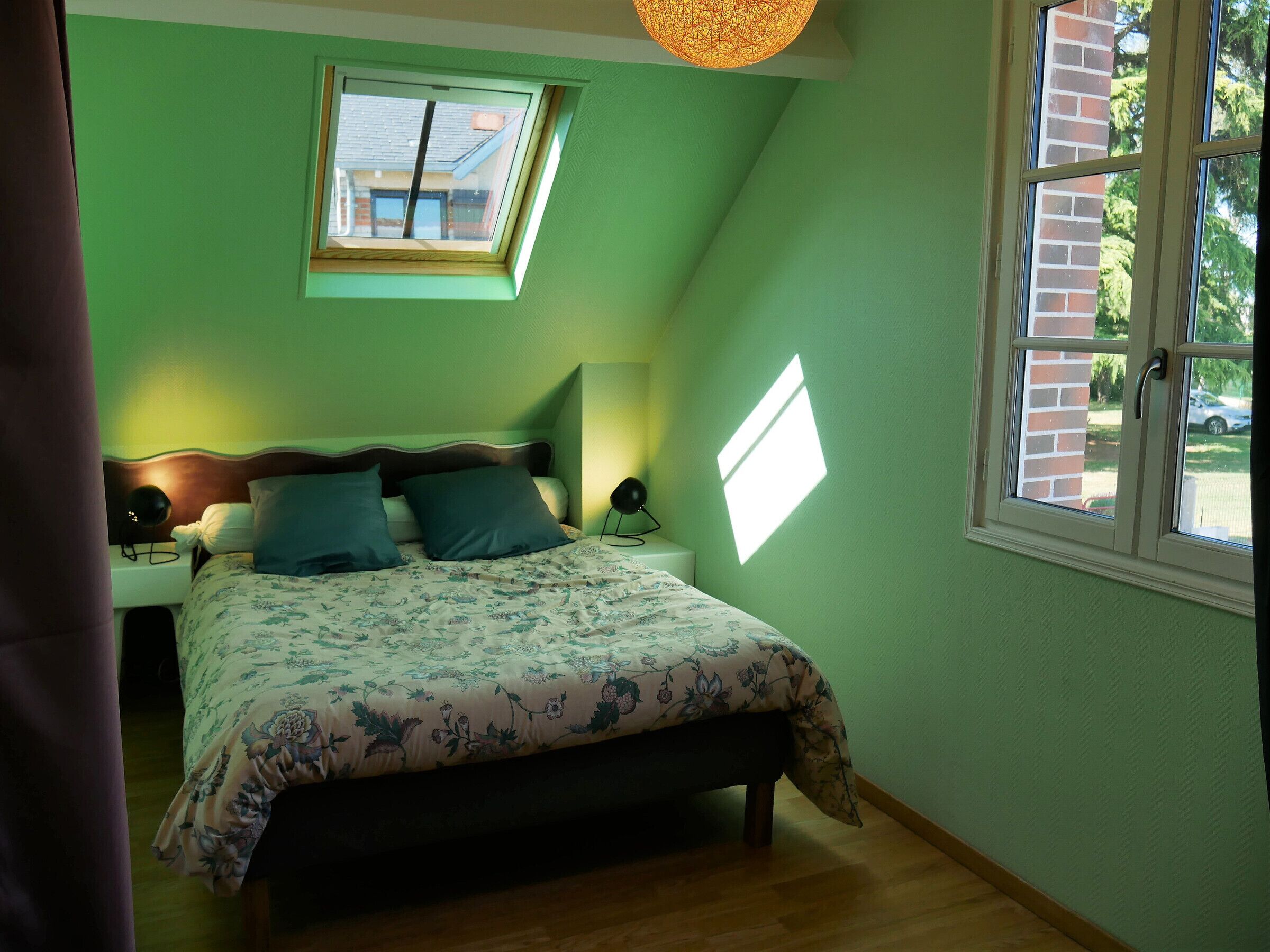 A bedroom with a bed, a nightstand, a bedside lamp, a window, and a skylight.