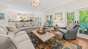 A spacious living room with a large sofa, a coffee table, and a grey armchair. There is a kitchen in the background with white cabinets and a dining area with a table and chairs.