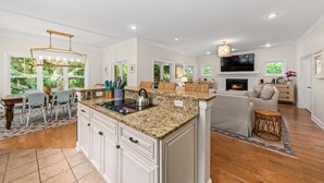 A spacious kitchen with granite countertops, white cabinetry, and a central island. There is a dining area with wicker chairs and a table, and a living room with a sofa and a fireplace.