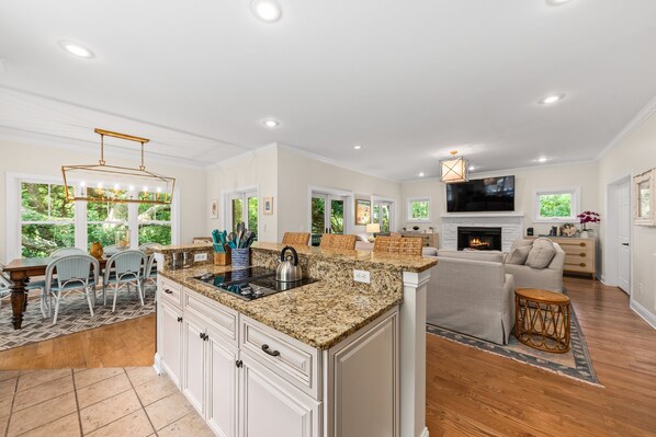 A spacious kitchen with granite countertops, white cabinetry, and a central island. There is a dining area with wicker chairs and a table, and a living room with a sofa and a fireplace.