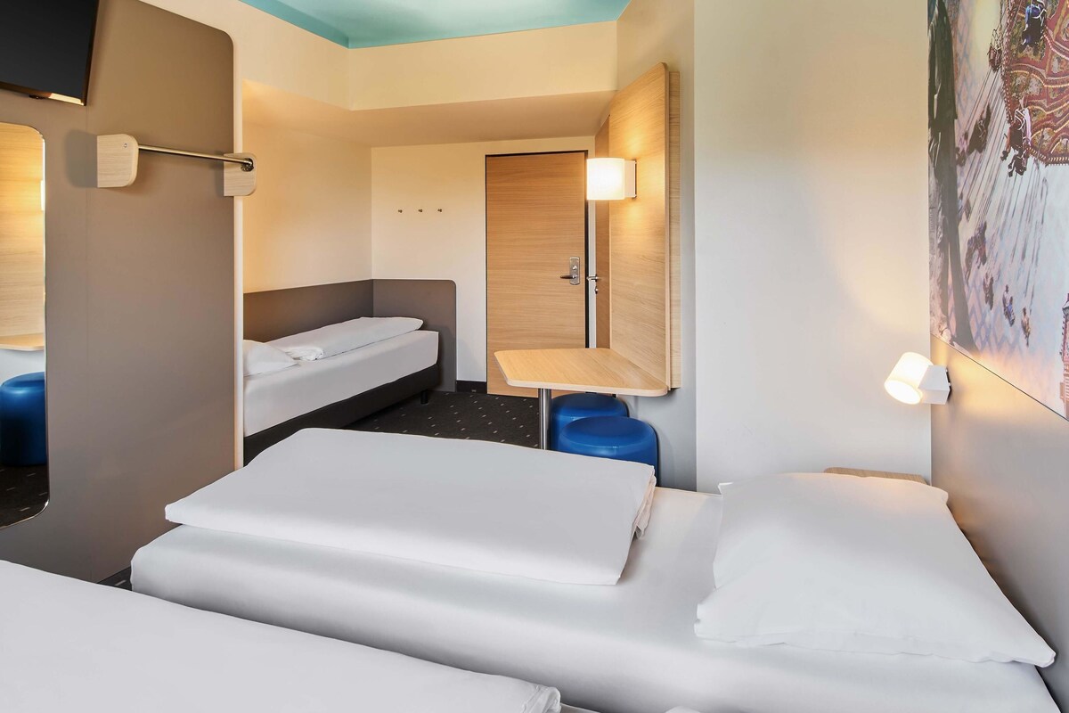 A hotel room with two beds, a small table, a chair, a wardrobe, and a TV.