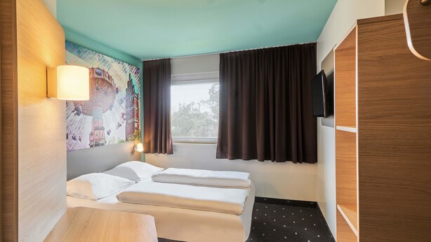 A compact hotel room with a bed, a small desk, a chair, a TV mounted on the wall, and a window with curtains. - B&B Hotel Darmstadt (Darmstadt)