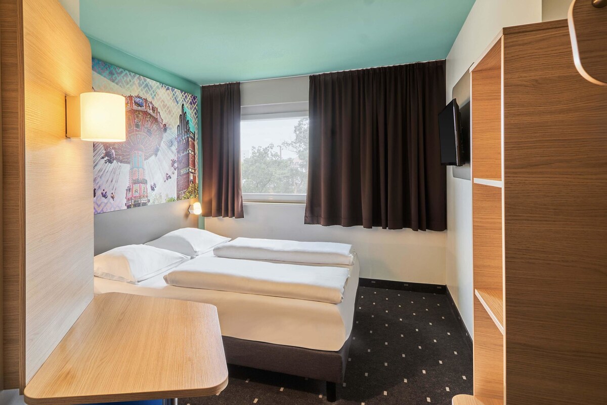 A compact hotel room with a bed, a small desk, a chair, a TV mounted on the wall, and a window with curtains.