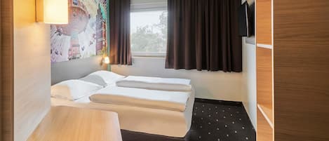 Habitación de hotel pequeña con cama, escritorio, silla, televisor montado en la pared y ventana con cortinas.