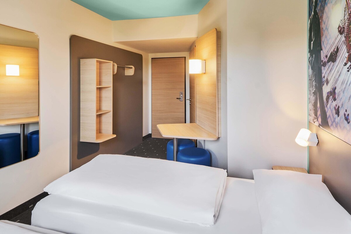 A hotel room with a bed, a desk, a chair, a wardrobe, and a wall-mounted shelf.