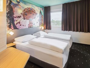 A hotel room with a bed, a nightstand, a desk, a wall-mounted lamp, a window with curtains, and a colorful mural on the wall. - B&B Hotel Darmstadt (Darmstadt)